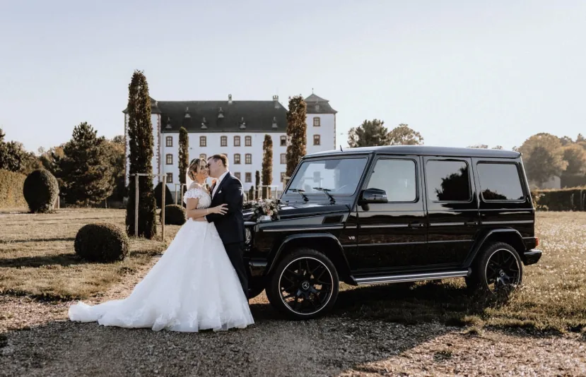 Brautpaar neben einer schwarzen Mercedes G-Klasse vor dem Schloss