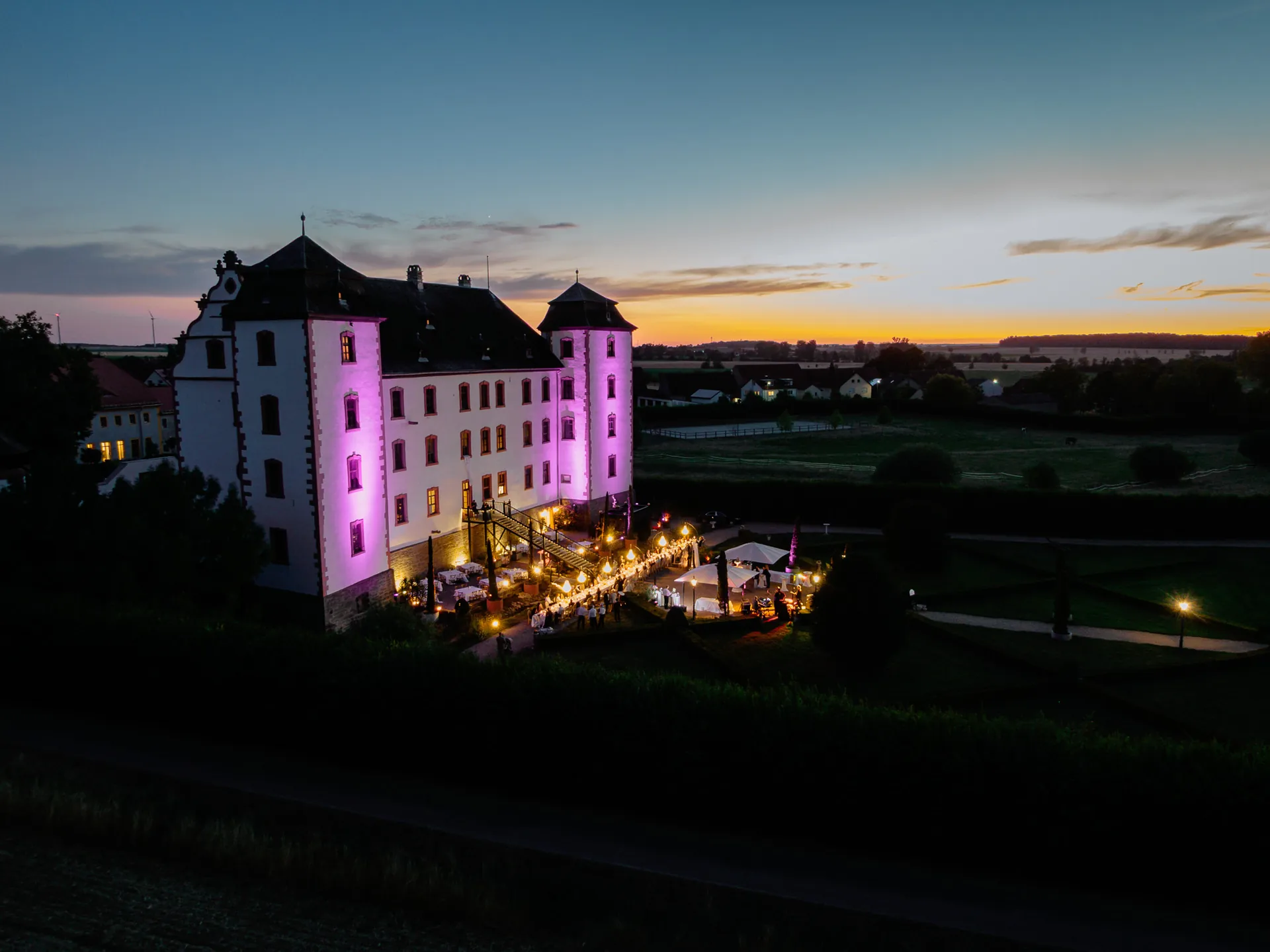 Violett angestrahltes Schloss mit beleuchteter Terrasse und Gästen in der blauen Stunde